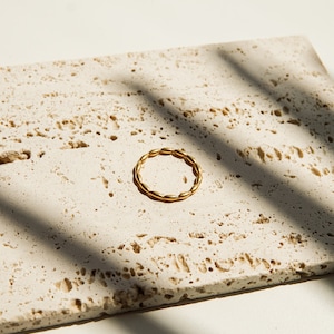 Może przedstawiać: Złoty pierścionek o skręconym wzorze, leżący na jasnobeżowej, teksturowanej powierzchni.