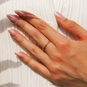 Op de afbeelding: Een close-up van een hand met een delicate gouden ring met een bloemenontwerp. De ring zit aan de ringvinger van de hand, die op een wit stofoppervlak rust. De nagels zijn gelakt met een lichtroze lak en hebben een witte punt.