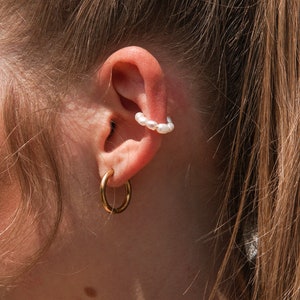 Könnte beinhalten: Nahaufnahme eines Ohrs mit zwei Ohrringen. Der obere Ohrring ist ein Perlenring und der untere Ohrring ist ein goldener Ring.