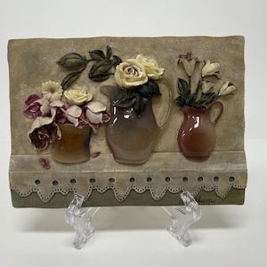 Peut inclure: Plaque murale décorative avec trois vases en céramique contenant des compositions florales. Les vases sont remplis de fleurs crème et bordeaux. La plaque a un fond neutre avec une bordure festonnée et est soutenue par un support transparent.