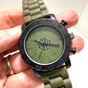 Puede incluir: Un reloj de pulsera negro con una correa y esfera verde oliva. La esfera del reloj muestra la hora con n&uacute;meros y agujas blancas. El reloj tiene un bisel negro con n&uacute;meros y una corona en el lateral.