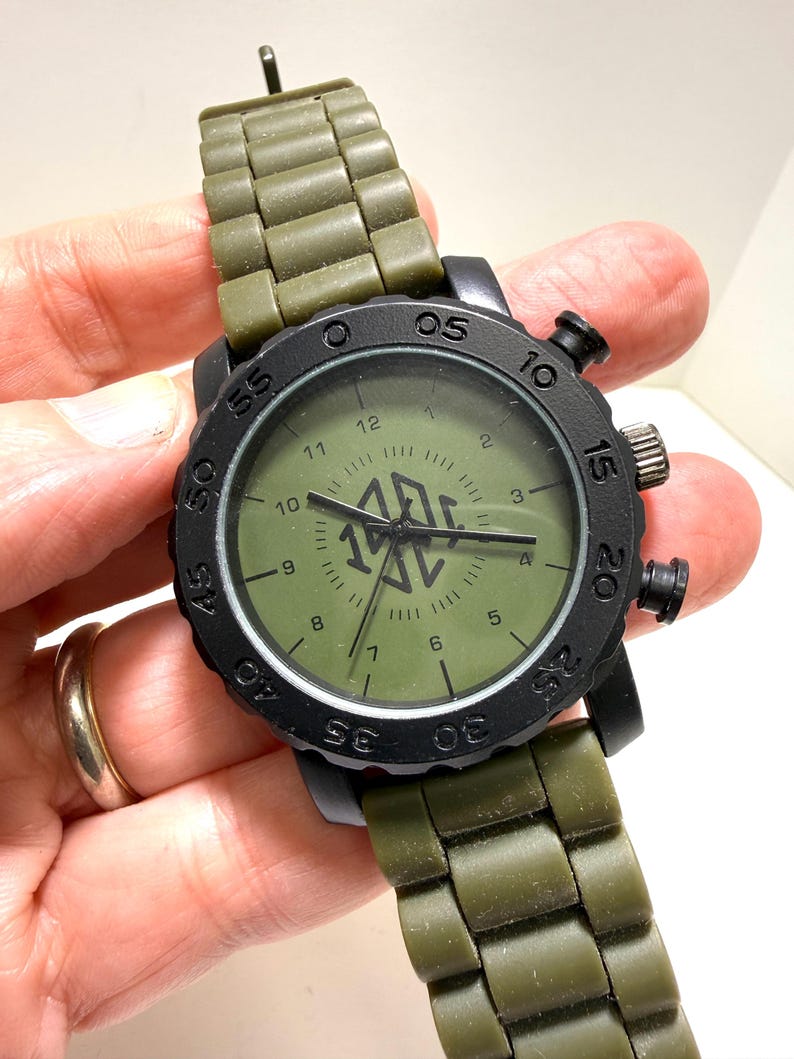 Puede incluir: Un reloj de pulsera negro con una correa y esfera verde oliva. La esfera del reloj muestra la hora con n&uacute;meros y agujas blancas. El reloj tiene un bisel negro con n&uacute;meros y una corona en el lateral.
