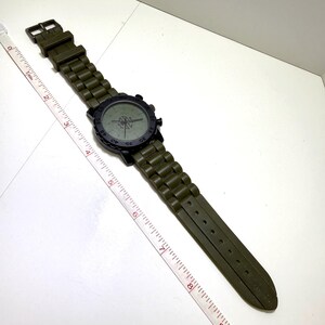 Puede incluir: Un reloj de pulsera verde oliva con un bisel negro y una correa verde oliva a juego. La esfera del reloj tiene un dise&ntilde;o de br&uacute;jula. La correa mide aproximadamente 23 cm de largo.