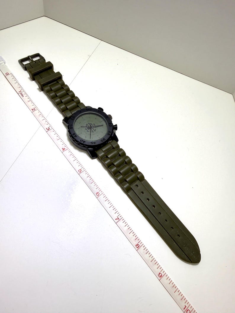 Puede incluir: Un reloj de pulsera verde oliva con un bisel negro y una correa verde oliva a juego. La esfera del reloj tiene un dise&ntilde;o de br&uacute;jula. La correa mide aproximadamente 23 cm de largo.