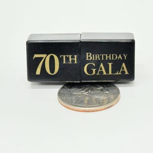 May include: A black box with gold lettering reading "70th Birthday GALA." A coin is placed below the box for size comparison. The box is likely a party favor or decoration for a 70th birthday celebration.