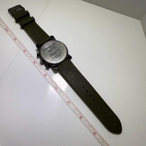 Puede incluir: Un reloj de pulsera verde oliva con esfera y correa negras. El reloj tiene una esfera redonda con m&uacute;ltiples esferas y el texto "COTTON ON" y "RN 129164". La correa del reloj mide aproximadamente 25 cm de largo.