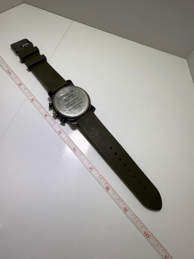 Puede incluir: Un reloj de pulsera verde oliva con esfera y correa negras. El reloj tiene una esfera redonda con m&uacute;ltiples esferas y el texto "COTTON ON" y "RN 129164". La correa del reloj mide aproximadamente 25 cm de largo.