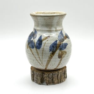 May include: A small, handcrafted ceramic vase with a cream-colored base and blue floral designs. The vase sits on a rustic, natural wood slice base. The vase has a rounded body and a slightly flared opening.
