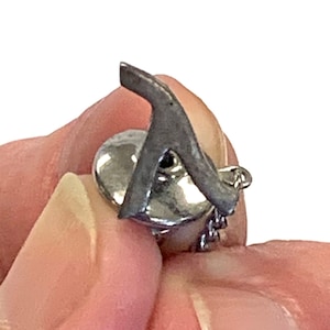 Może przedstawiać: Srebrna spinka do kołnierzyka z ozdobnym, stylizowanym literą A i przymocowaną do niej łańcuszkiem. Spinka jest trzymana w dłoni.