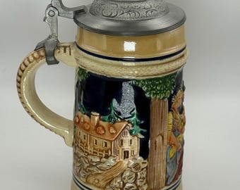 Puede incluir: Una jarra de cerveza de cerámica con tapa y asa metálicas con bisagras. La jarra presenta una escena en relieve detallada de un edificio y figuras en un bosque, con una combinación de colores crema y azul. La jarra mide aproximadamente 20 cm de altura.