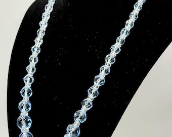 May include: A light blue beaded necklace with faceted beads of varying sizes. The necklace is displayed on a black mannequin bust, showcasing its length and design. The beads have a clear, sparkling appearance.