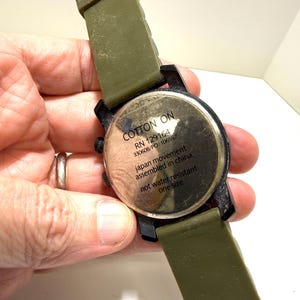 Puede incluir: Primer plano de un reloj de pulsera con una correa verde oliva y una esfera negra. La esfera del reloj tiene el texto "COTTON ON" y otros detalles. El reloj no es resistente al agua y es de talla &uacute;nica.
