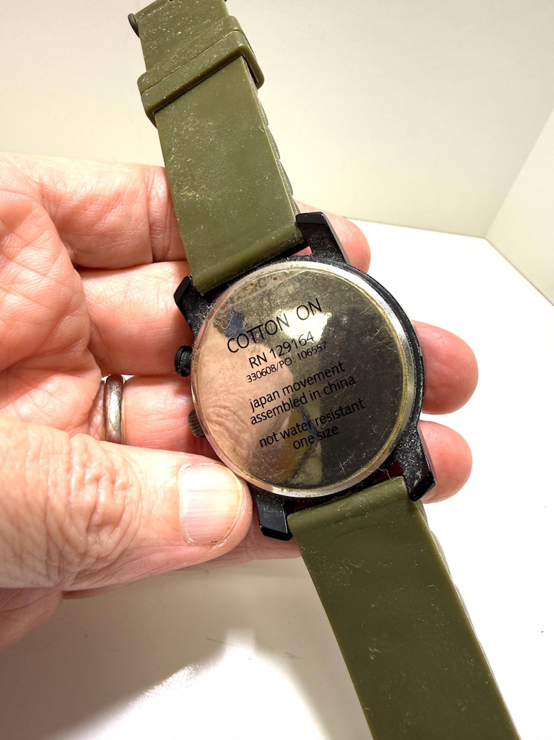 Puede incluir: Primer plano de un reloj de pulsera con una correa verde oliva y una esfera negra. La esfera del reloj tiene el texto "COTTON ON" y otros detalles. El reloj no es resistente al agua y es de talla &uacute;nica.
