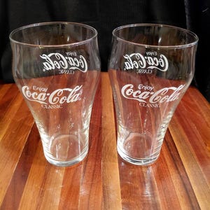 以下が含まれることがあります： 前面に白いCoca-Cola ClassicのロゴとEnjoy Coca-Colaの文字がプリントされた、2つの透明なグラス。グラスは飲み口が広く底が狭いテーパー状の形状で、冷たい飲み物を注ぐために設計されています。