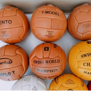 Puede incluir: Una colección de balones de fútbol vintage en varios tonos de marrón y amarillo. Cada balón presenta paneles cosidos y texto impreso, incluyendo "TIENTO", "T-MODEL" y "SWISS WORLD CHAMPION". Los balones están dispuestos juntos, mostrando sus diseños únicos.
