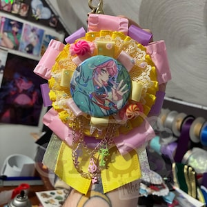 May include: A decorative rosette badge featuring a central circular pinback button with an anime character illustration, surrounded by layers of pleated pink and yellow ribbons, white lace, small candy-shaped charms, and dangling metallic key charms.