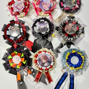 Puede incluir: Nueve insignias decorativas de roseta con varios diseños. Cada insignia presenta una imagen central, rodeada de capas de cintas en colores como rojo, rosa, negro y blanco. Algunas tienen cadenas y detalles colgantes.