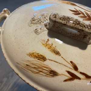 May include: A white ceramic serving dish with a brown handle and a floral design featuring wheat stalks. The dish has a speckled glaze and a rustic look.
