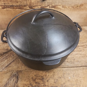 May include: A black cast iron Dutch oven with a lid and handle. The pot has a rounded shape and sits on a wooden surface. The lid has a handle and a wire handle. The pot is designed for cooking.