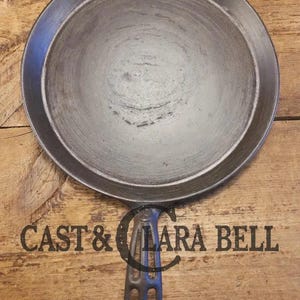 May include: A round, dark gray cast iron pan with a long handle. The pan has a slightly textured surface and a raised edge. The words "CAST & LARA BELL" are printed on the wooden surface below the pan.