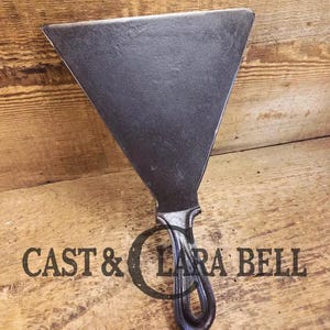 May include: A dark gray, triangular cast iron spatula with a curved handle and a loop for hanging. The text "CAST & LARA BELL" is printed on the handle. The spatula is set against a wooden background.