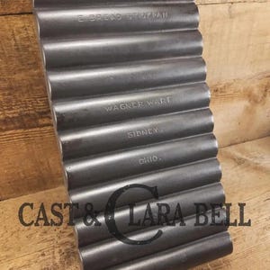 May include: A dark gray cast iron bread stick pan with a ribbed design. The pan has the words "E BREAD STICK PAN" and "WAGNER WARE SIDNEY, OHIO" embossed on it. The pan is rectangular with a handle and is set against a wooden background.