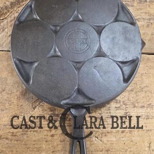 May include: A cast iron pan with seven circular molds, perfect for making pancakes or small cakes. The pan has a handle and the text "CAST & LARA BELL" is printed on the surface. The pan is dark gray and sits on a wooden surface.