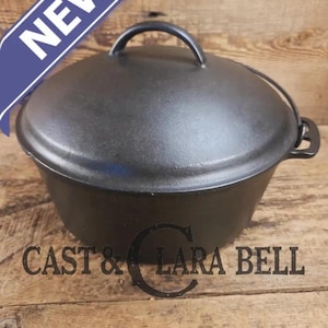 May include: A black cast iron Dutch oven with a lid and handle. The word "NEW" is displayed on a blue banner. The text "CAST & LARA BELL" is printed on the wooden surface.