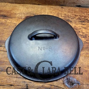 May include: Black cast iron lid with a handle and the text "No. 8" on the top. The lid is sitting on a wooden surface.
