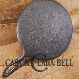 May include: A black cast iron skillet with a textured surface and a handle. The skillet is round and has the text "CAST & LARA BELL" printed on it. The skillet is on a wooden surface.