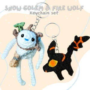 May include: A set of two crocheted keychains. One keychain is a white snow golem with brown accents and a green leaf on its head. The other keychain is a black fire wolf with orange accents. The keychains are attached to silver key rings.