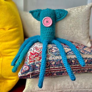 May include: A handmade teal crochet octopus with a pink button eye and long tentacles. The octopus has two small ear-like appendages. It is resting on a patterned cushion with a yellow cushion in the background.