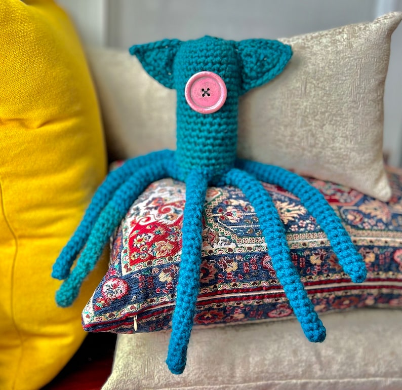 Puede incluir: Un pulpo de ganchillo turquesa hecho a mano con un ojo de bot&oacute;n rosa y largos tent&aacute;culos. El pulpo tiene dos peque&ntilde;os ap&eacute;ndices en forma de oreja. Est&aacute; sobre una almohada estampada.