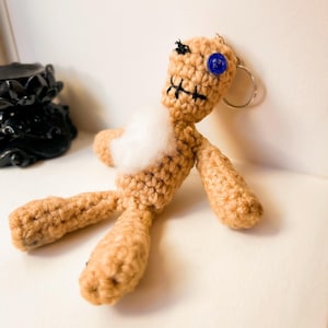 Puede incluir: Una muñeca de crochet con un ojo de botón azul, boca cosida y relleno blanco. La muñeca está tumbada de espaldas con los brazos extendidos y las piernas dobladas. La muñeca tiene un llavero plateado sujeto a su cabeza.