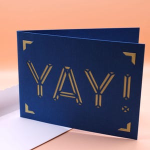May include: A blue card with a gold cutout of the word "YAY!" in a geometric font.