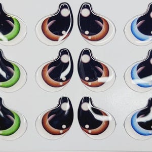 May include: A sheet of 12 pairs of plastic doll eyes. The eyes are black with white pupils and have different colored accents around the pupils. The accents are green, brown, and blue.