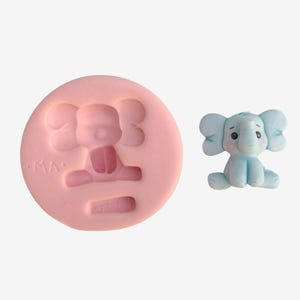May include: A light blue elephant figurine next to a pink silicone mold. The elephant has large ears, a trunk, and is sitting down. The mold is round with an elephant shape and the letters "MA" on the side.