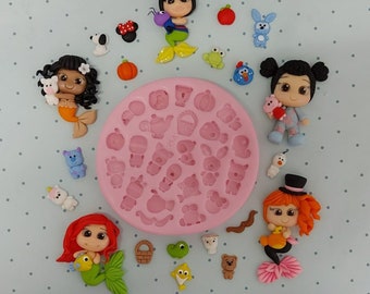 Puede incluir: Un molde de silicona rosa con varias formas, incluyendo una tetera, una cesta, un pez y animales. Alrededor del molde hay varias figuritas de arcilla polimérica hechas a mano, incluyendo sirenas, una niña en pijama y una niña con un sombrero de copa.