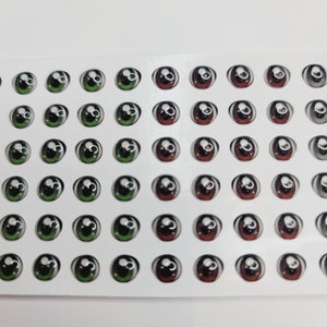 Könnte beinhalten: Eine Plastikfolie mit 48 runden, selbstklebenden Glitzern. Die Augen sind in 6 Reihen zu je 8 angeordnet. Die Augen sind grün, braun und schwarz mit weißen Pupillen.