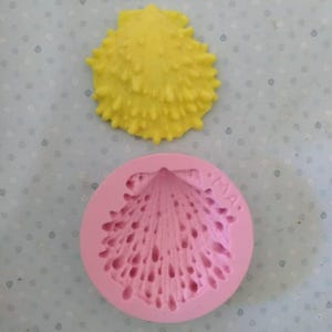 May include: A pink silicone mold for making a yellow seashell cake decoration. The mold has a detailed design of a seashell with a textured surface. The mold is labeled "MIA."