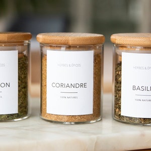 Peut inclure: Trois bocaux en verre avec des couvercles en bambou, chacun contenant des herbes séchées et étiqueté avec le nom de l'herbe en français. Les étiquettes indiquent également "Herbes & Epices" et "100% Naturel". Les herbes sont l'estragon, la coriandre et le basilic.