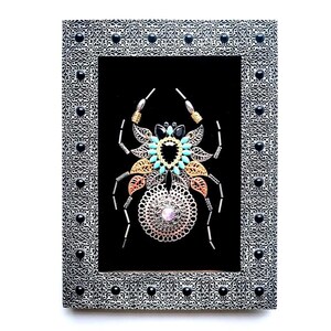May include: A framed artwork with a decorative spider design. The spider is made from silver, gold, and copper metals, and decorated with turquoise and black gemstones. The silver frame has black accents.