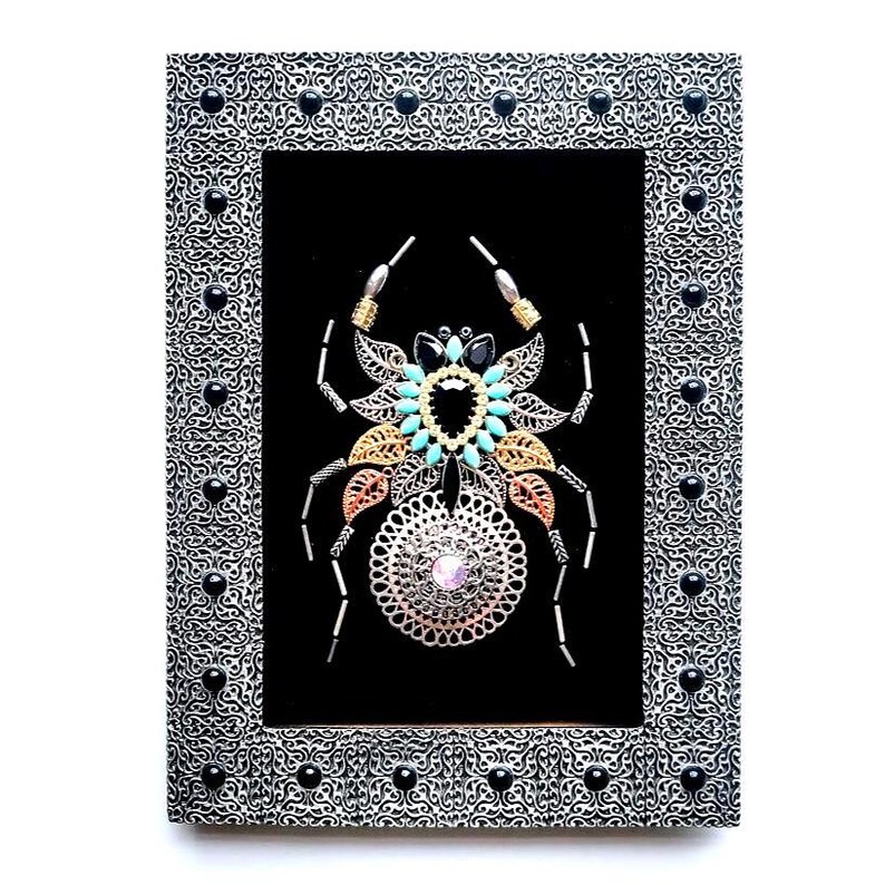 May include: A framed artwork with a decorative spider design. The spider is made from silver, gold, and copper metals, and decorated with turquoise and black gemstones. The silver frame has black accents.