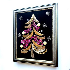 Op de afbeelding: Ingelijst kunstwerk met een kerstboomontwerp. De boom is samengesteld uit goudkleurige bananenvormen, roze edelstenen en gouden bladaccenten, tegen een zwarte achtergrond. Zilveren sneeuwvlokken en ornamenten maken de feestelijke look compleet.