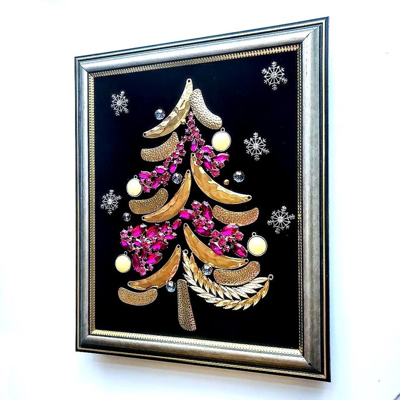Op de afbeelding: Ingelijst kunstwerk met een kerstboomontwerp. De boom is samengesteld uit goudkleurige bananenvormen, roze edelstenen en gouden bladaccenten, tegen een zwarte achtergrond. Zilveren sneeuwvlokken en ornamenten maken de feestelijke look compleet.