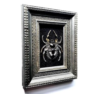 May include: A framed artwork featuring a detailed spider design. The spider is crafted from various metallic elements, including silver and gold tones, and features a large, dark gemstone at its center. The artwork is set against a dark background within a silver frame.