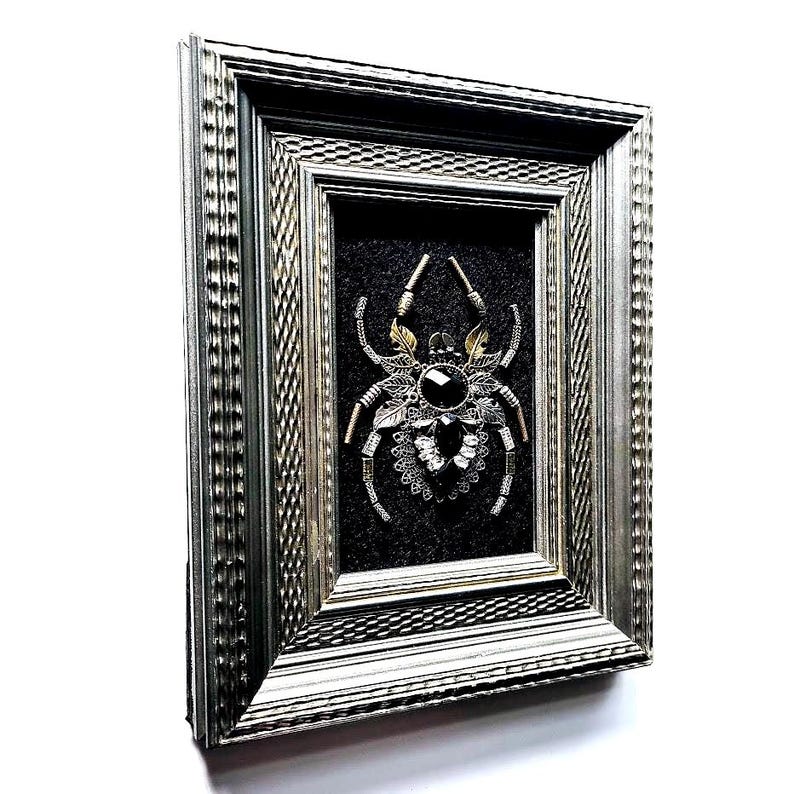 May include: A framed artwork featuring a detailed spider design. The spider is crafted from various metallic elements, including silver and gold tones, and features a large, dark gemstone at its center. The artwork is set against a dark background within a silver frame.