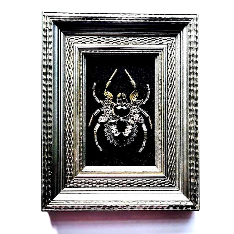 May include: Framed artwork featuring a detailed spider design. The spider is crafted with gold and silver-toned metal, adorned with a large black gem at its center. The artwork is set against a black background within a silver-colored frame.