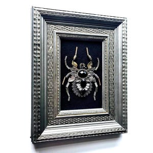May include: Framed artwork featuring a detailed spider design. The spider is crafted from metallic elements, including gold and silver tones, with a large black gem at its center. The artwork is set against a black background within a silver frame.