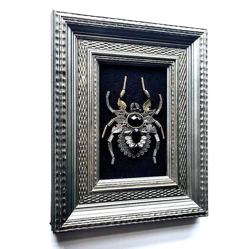 May include: Framed artwork featuring a detailed spider design. The spider is crafted from metallic elements, including gold and silver tones, with a large black gem at its center. The artwork is set against a black background within a silver frame.